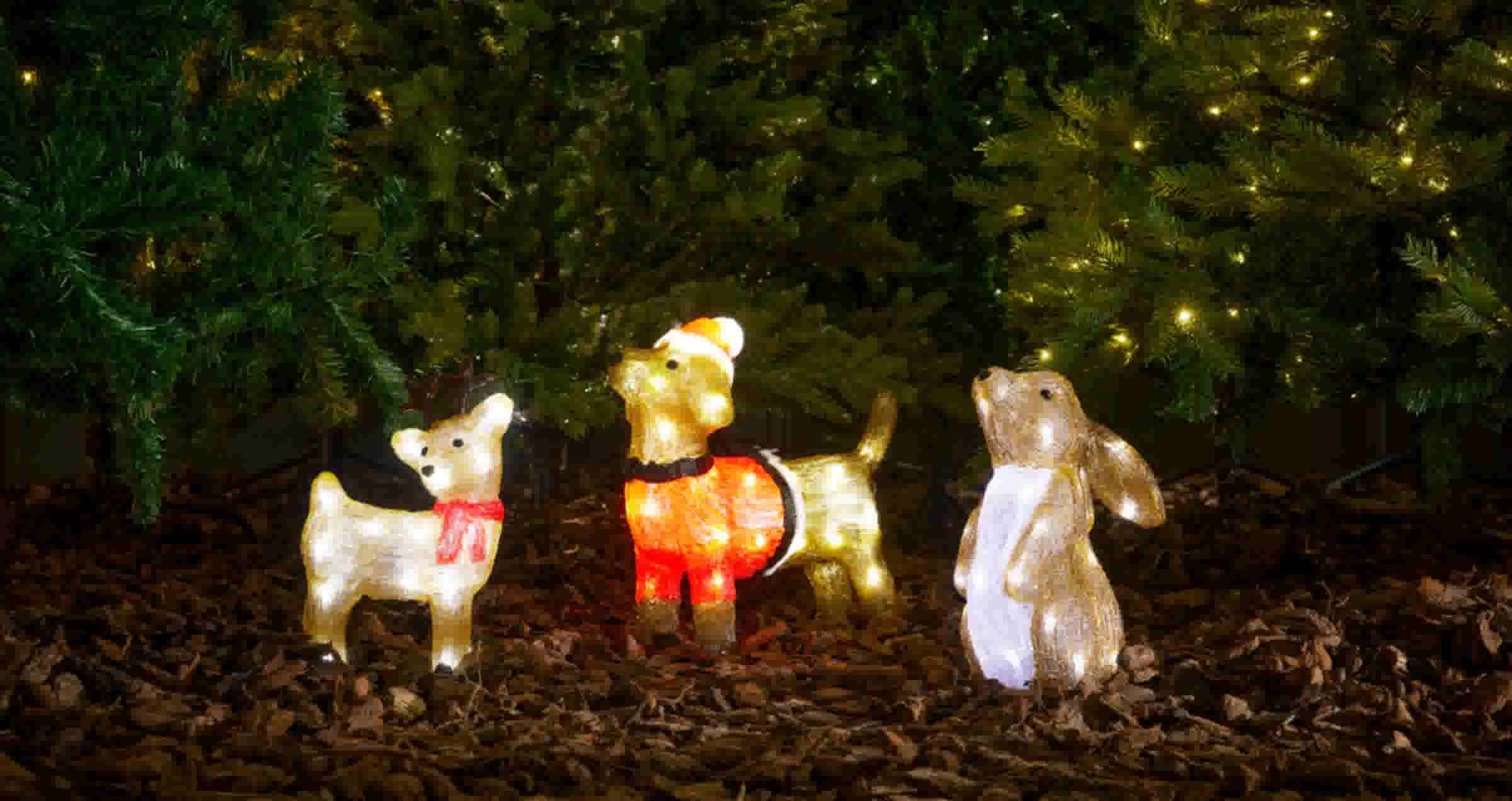 Verlichte kerstfiguren onder dennenbomen: een wit hertje met rode sjaal, een rendier met kerstmuts en een witte konijn.