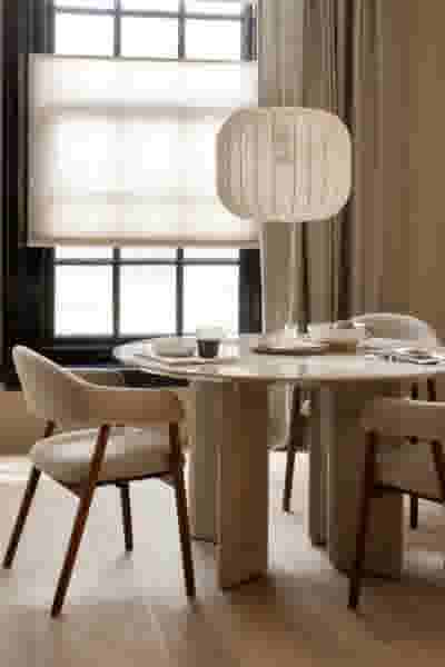 Minimalistische eetkamer met ronde beige tafel, stoelen met houten poten en een witte hanglamp. Lichte tinten en groot raam op de achtergrond.