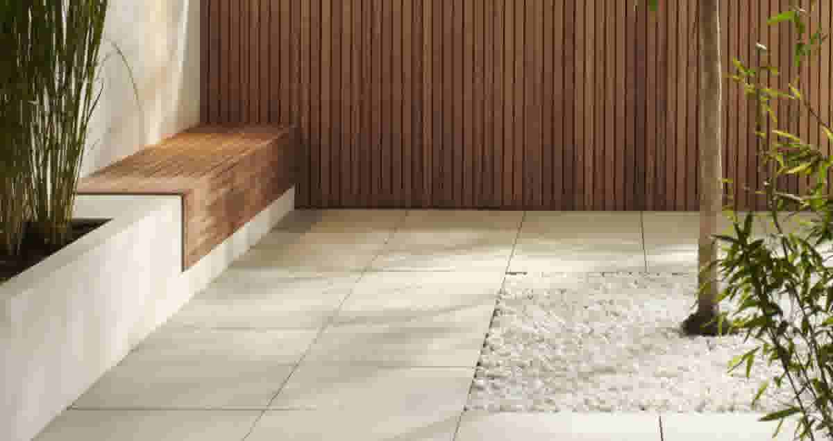Moderne patio met lichte tegels, houten zitbank en wandbekleding, witte kiezelstenen rond een bamboeplant en groen.