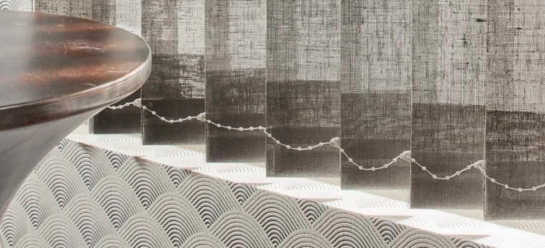 Grijze verticale jaloezieën met decoratieve koordverbinding, naast een houten tafelblad en een kussen met golfpatroon.