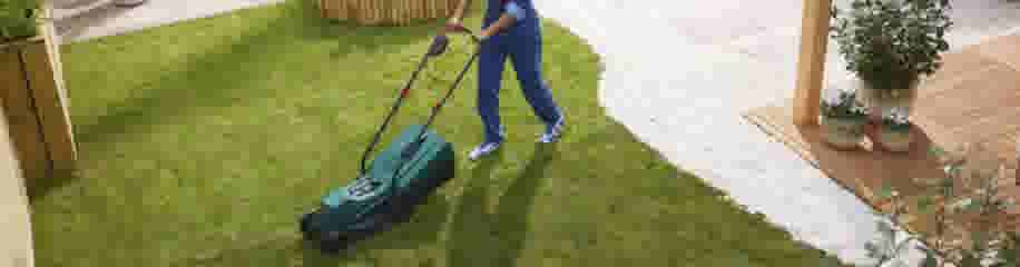 Persoon in blauwe kleding maait het gazon met een groene grasmaaier in een verzorgde tuin met houten terras.