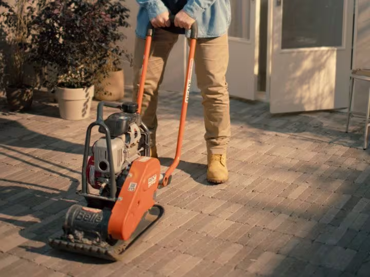 Persoon bedient een gehuurde oranje trilplaat op een terras van bakstenen, gekleed in beige broek en blauw shirt.