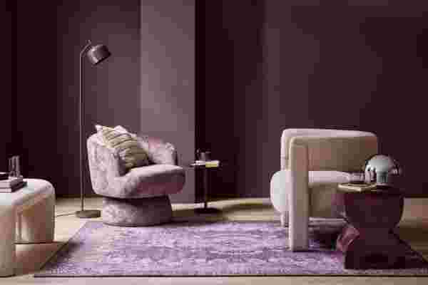 Modern woonkamer met paarse muren, roze fauteuil, beige stoel, bijzettafels en een paars tapijt onder stijlvolle verlichting.