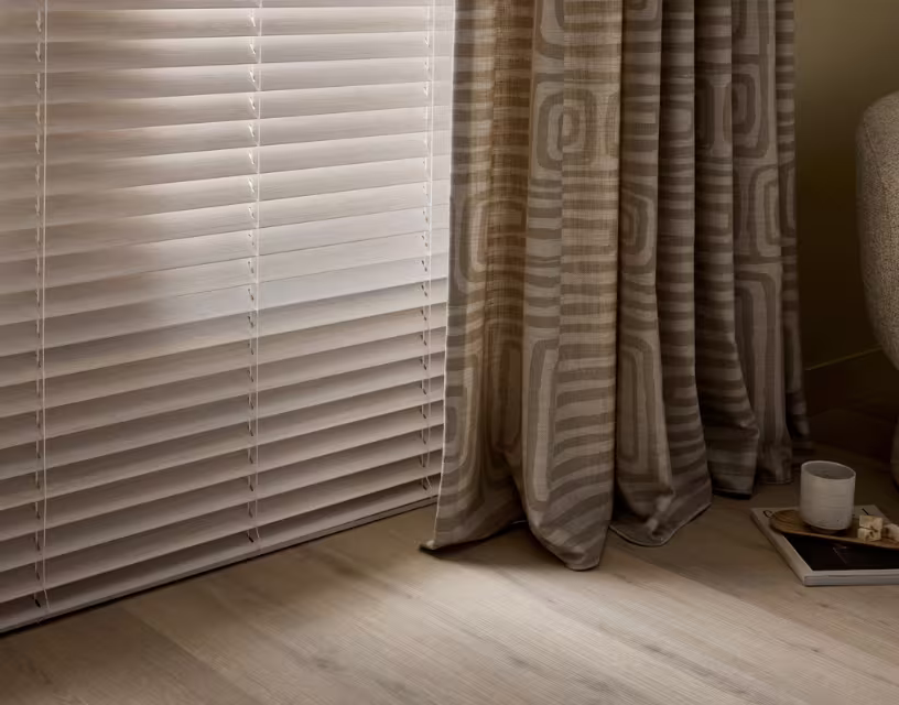 Witte horizontale jaloezieën naast beige gordijnen met geometrisch patroon in een moderne woonkamer met houten vloer.