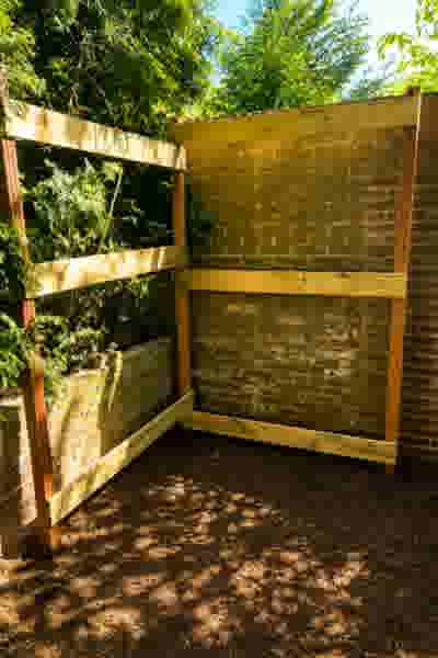 Houten frame tegen een bakstenen muur in een tuin, omgeven door groen gebladerte en bruine grond met schaduwen.