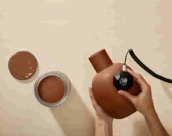 Hand scant met een kleurscanner een terracotta vaas. Daarnaast staat een open verfpot op een beige ondergrond.