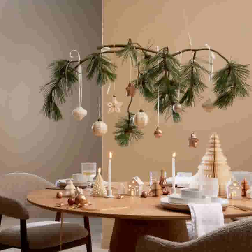 Feestelijke eettafel met hangende dennentakken versierd met kerstballen en sterren, papieren kerstbomen en brandende kaarsen.