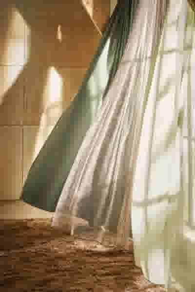 Lichtdoorlatende groene en witte gordijnen wapperen zachtjes voor een houten wand, met zonlicht dat door het raam valt.