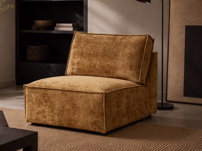 Okergele modulaire fauteuil zonder armleuningen op een juten vloerkleed, met donkere boekenkast en decoratieve elementen op de achtergrond.