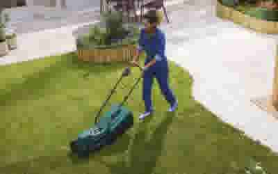 Persoon in blauwe kleding maait het gras met een elektrische grasmaaier in een tuin met verhoogde plantenbakken.