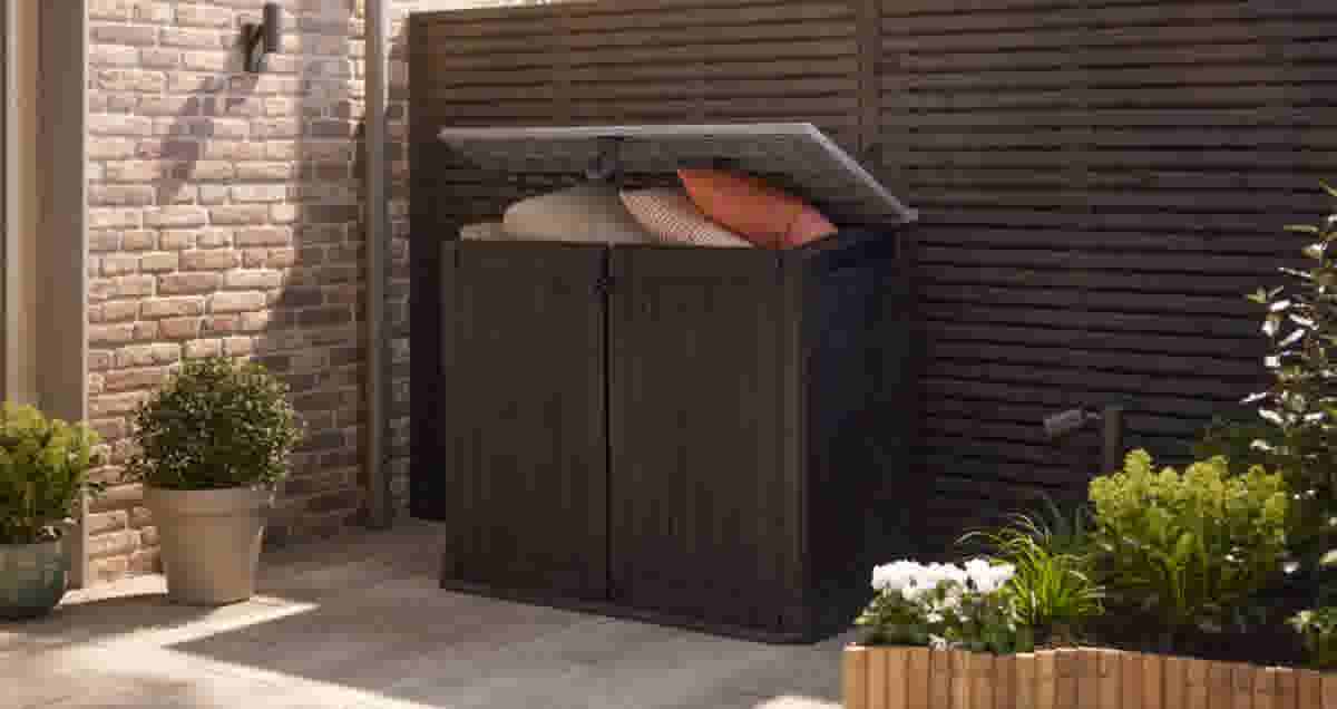 Donkere tuinkast met open deksel, gevuld met kussens, op een patio naast een bakstenen muur en groene planten.