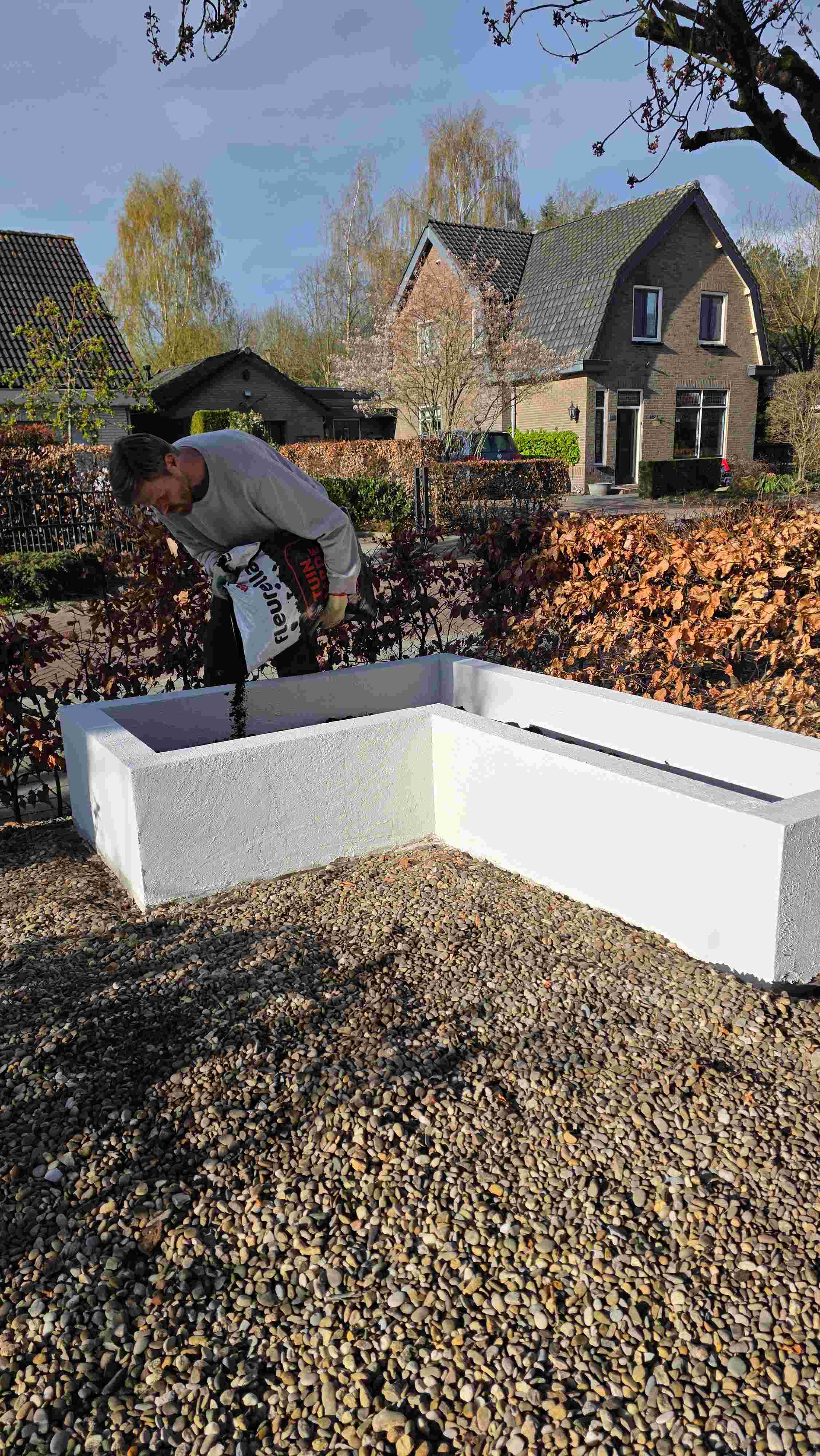 Persoon werkt aan witte verhoogde plantenbak in tuin met kiezelstenen, op achtergrond bakstenen huizen en bloeiende bomen.