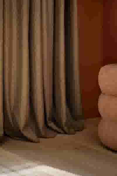 Bruine puddle gordijnen die op de houten vloer hangen naast een lichtroze gestoffeerd meubel in een warme, minimalistische ruimte.