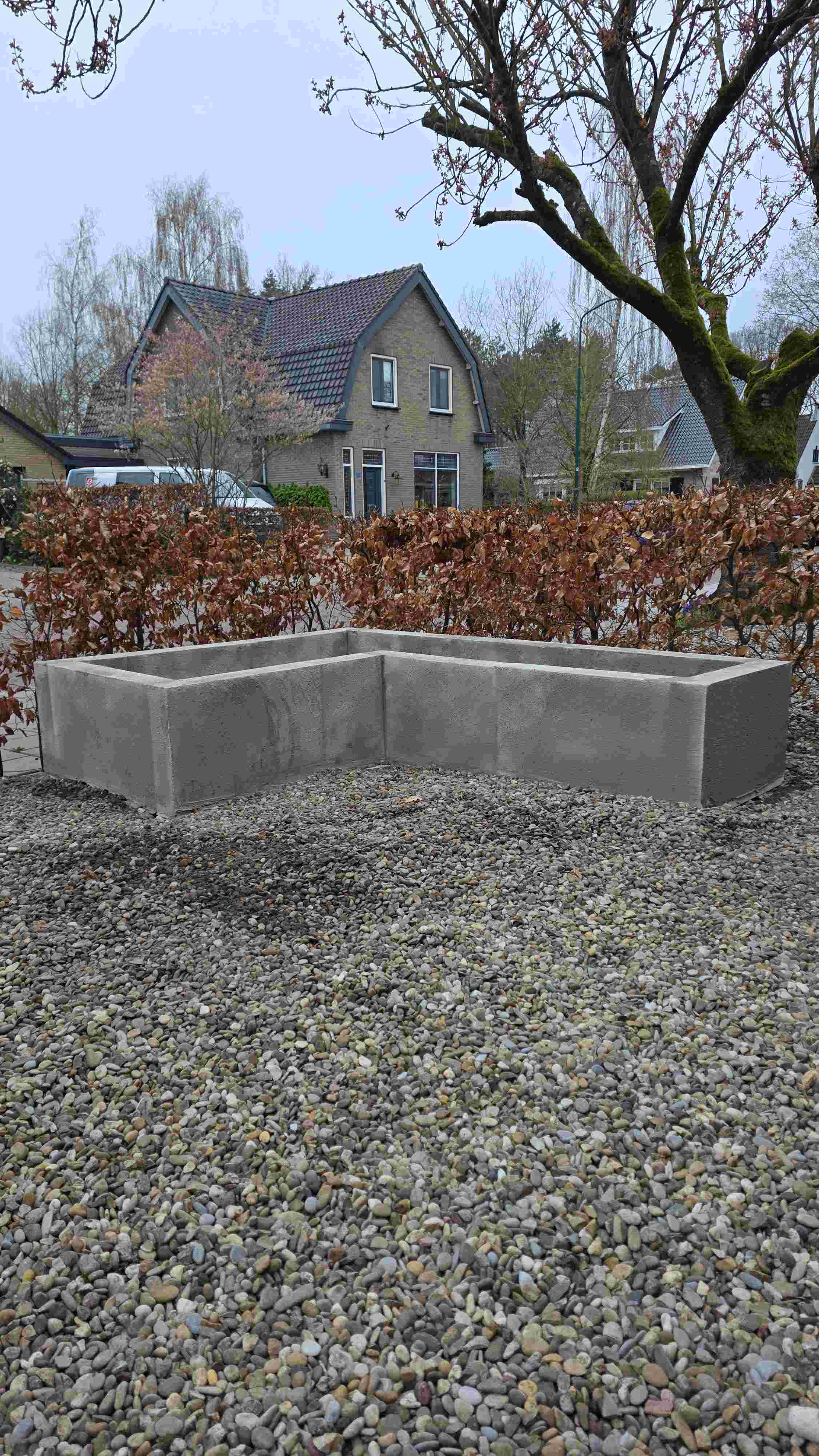 L-vormige betonnen plantenbak op grindondergrond met bruine haag en bakstenen woning op de achtergrond onder bewolkte hemel.