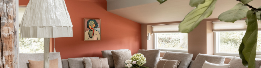 Moderne woonkamer met oranje muur, beige bank, witte hanglamp en kunstwerk boven de bank met grote ramen.