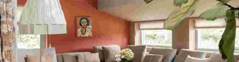 Moderne woonkamer met oranje muur, beige bank, witte hanglamp en kunstwerk boven de bank met grote ramen.