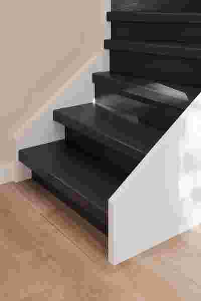Donkere houten trap met witte zijkant op een lichte houten vloer, tegen een beige muur in een moderne woning.