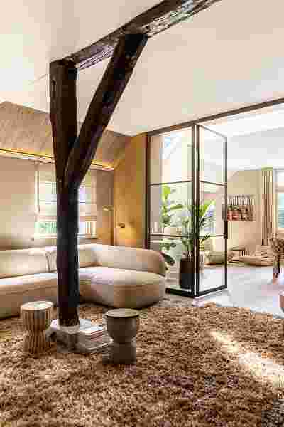 Moderne woonkamer met donkere houten balken, beige gebogen bank, glazen serrekast met plant en bruin hoogpolig tapijt.