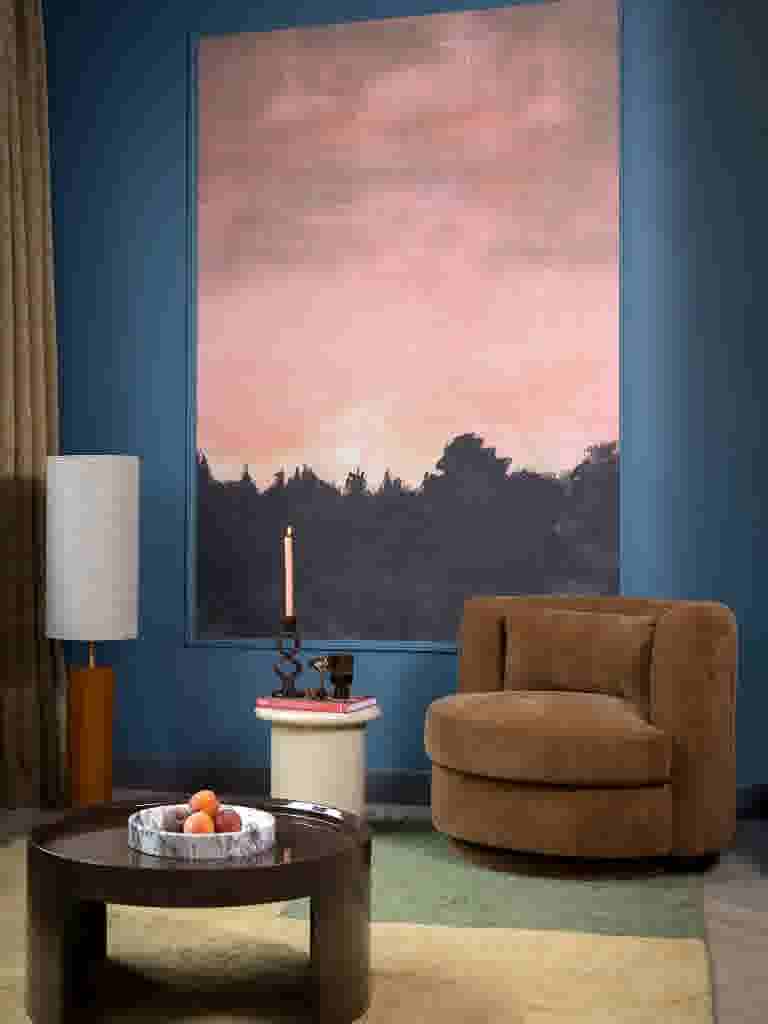 Sfeerbeeld van een woonkamer in blauwtinten met een bruine fauteuil, een ronde bijzettafel met kaars en boek, een lage salontafel met schaal fruit en een vloerlamp, voor een groot schilderij van een zonsondergang.