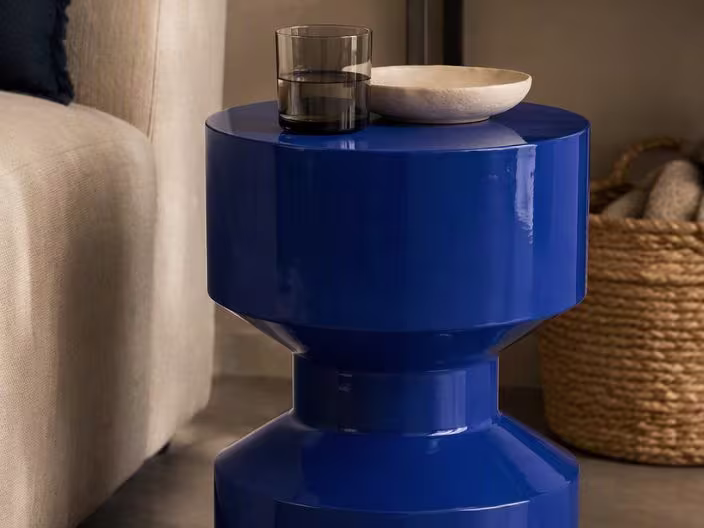 Glanzend kobaltblauwe bijzettafel met een rookglas en beige schaaltje erop, naast een beige bank en rieten mand.