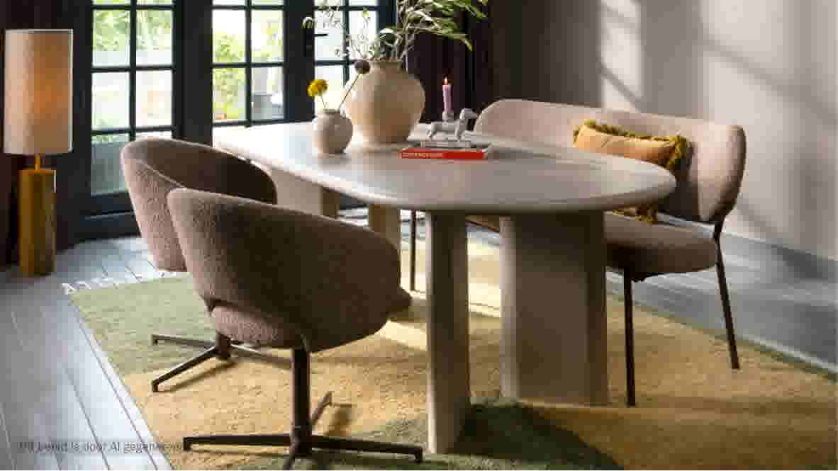 Sfeerbeeld van een eetkamer met ovale tafel, gestoffeerde stoelen en bank, vazen met bloemen, een kandelaar met teckelbeeldje en het boek 'Super Nice Stuff' van Theo-Bert Pot, op een groen-geel vloerkleed.