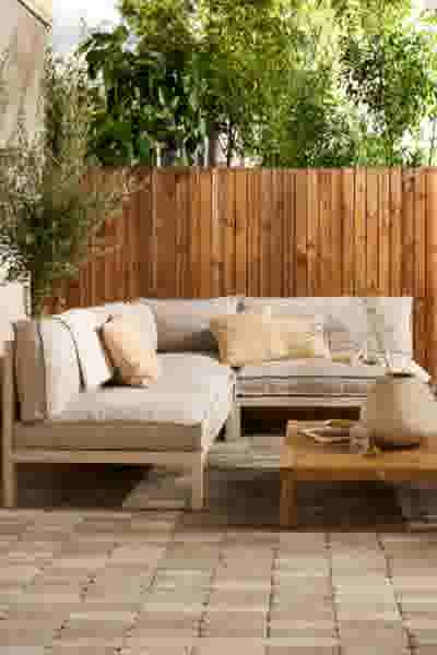 Beige hoekbank met kussens op een terras, houten salontafel, ronde vaas en houten schutting met groene planten op de achtergrond.