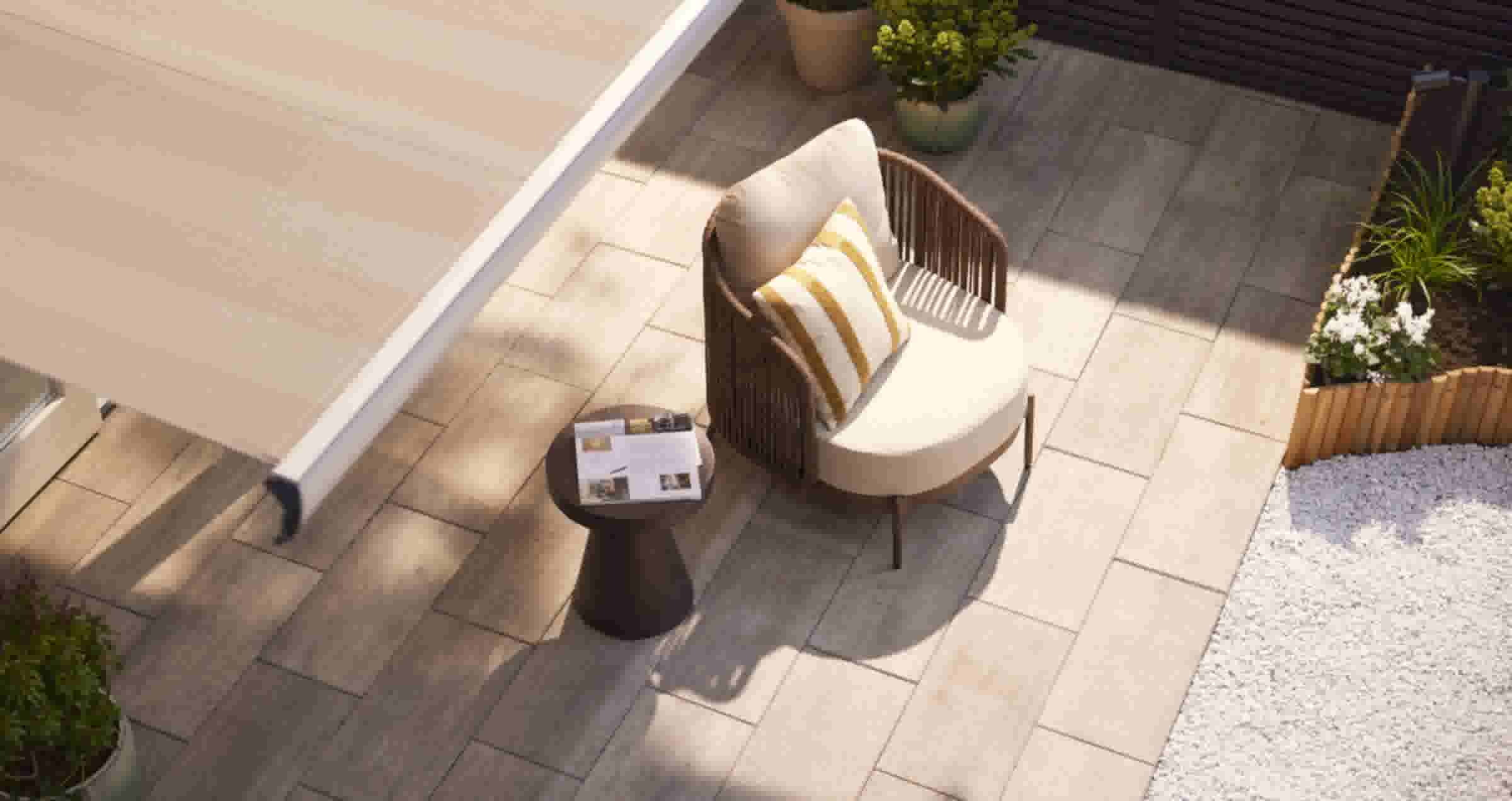 Bovenaanzicht van een terras met beige tegels, een rotan stoel met kussen en een donker bijzettafeltje met tijdschrift. Planten op de achtergrond.