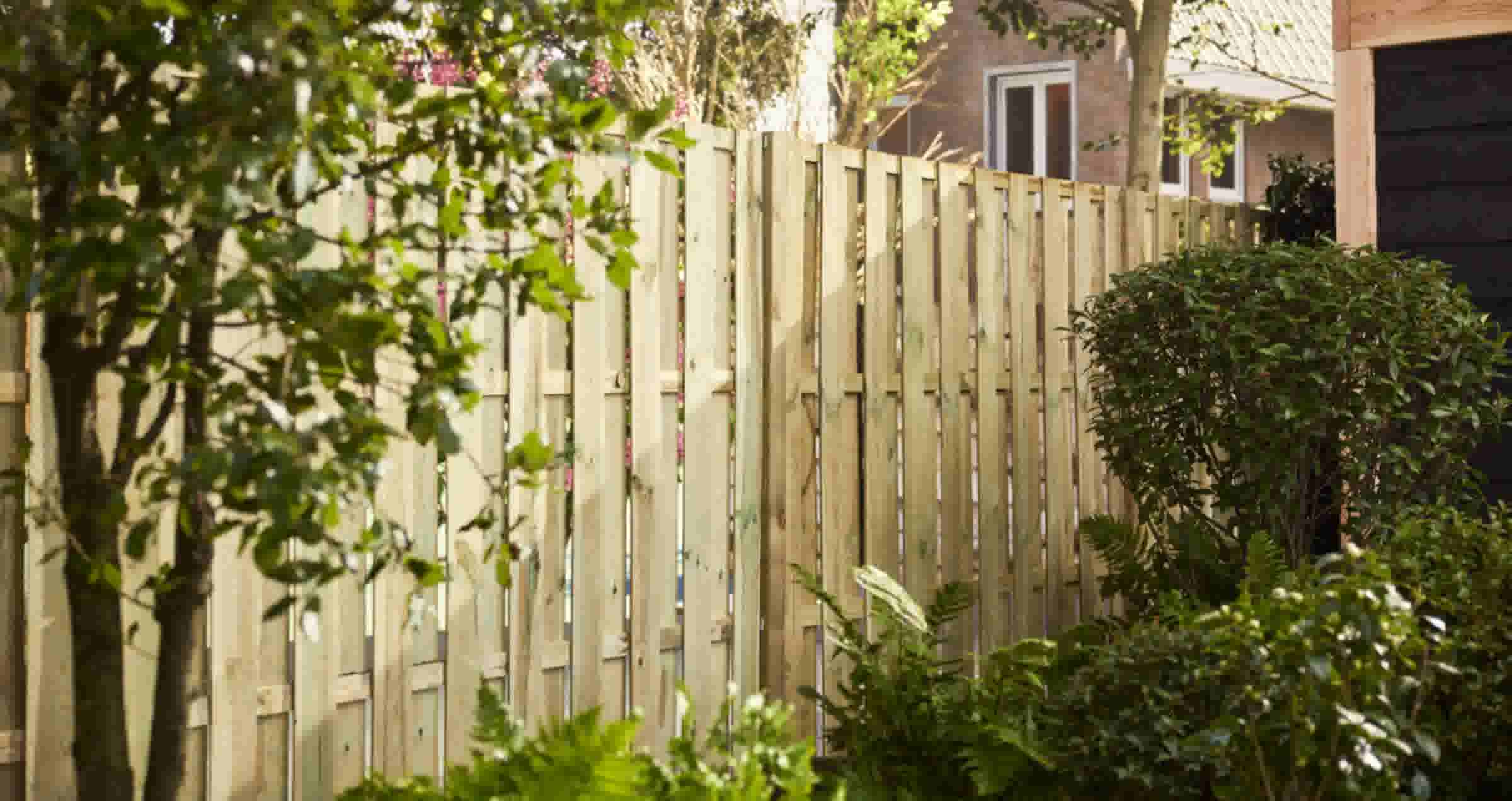 Houten verticale schutting in een tuin omgeven door groene planten en bomen, met een bakstenen huis op de achtergrond.