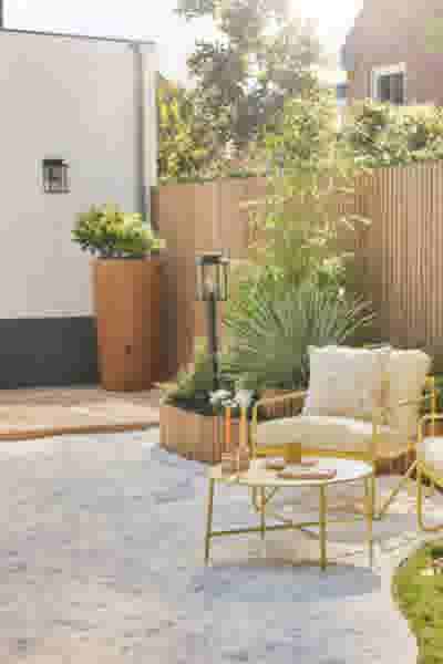 Zonnige tuin met gele rotan stoelen en bijzettafel op grind, houten schutting, grote terracotta pot en weelderige planten op de achtergrond.