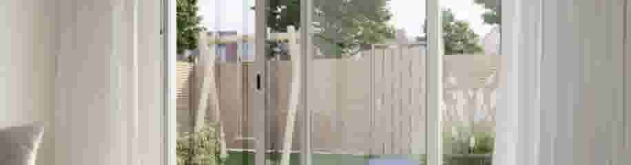 Uitzicht vanuit een woonkamer door grote glazen schuifdeuren met een deurhor naar een tuin met houten schutting en groene planten.