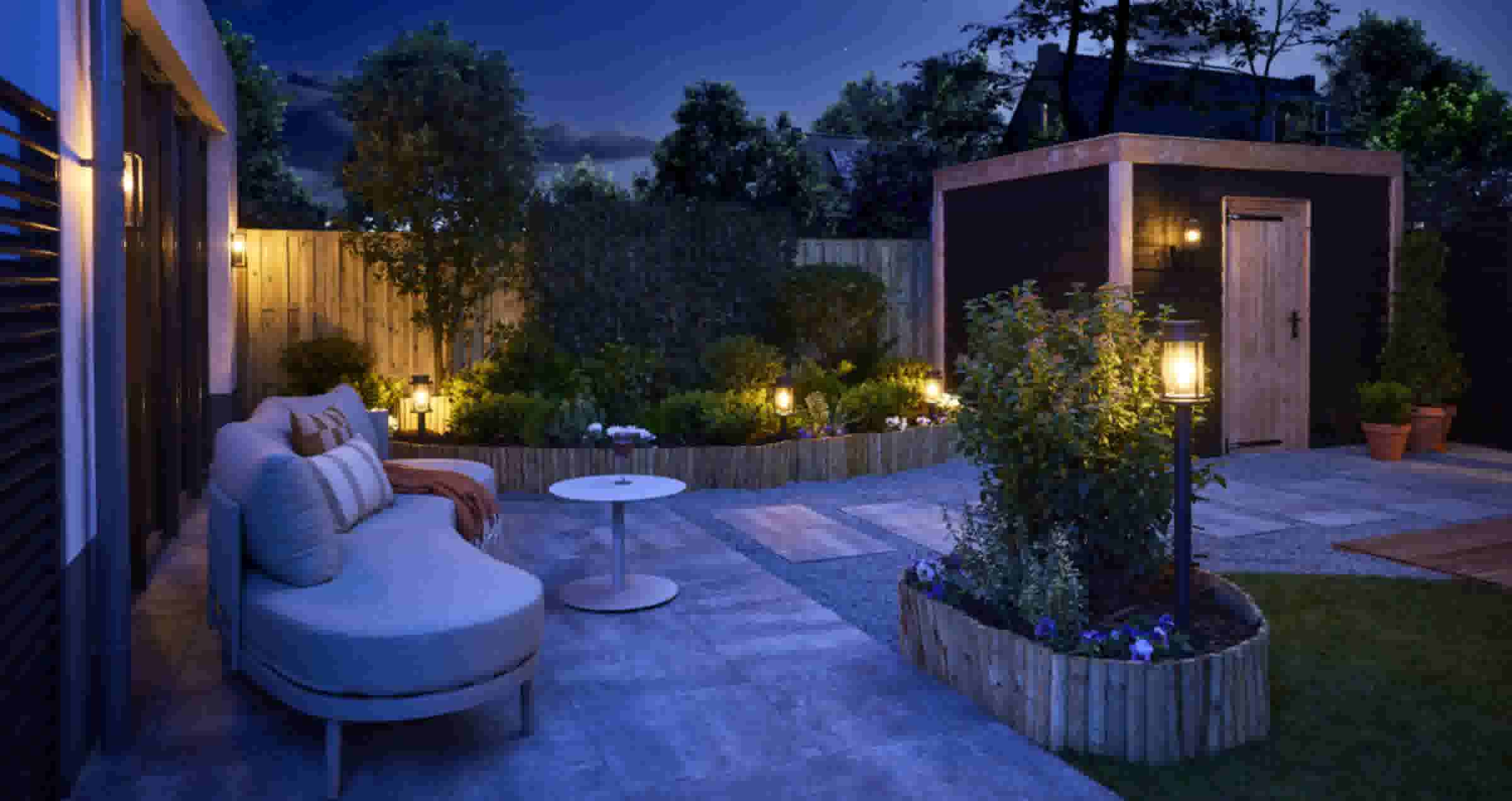 Sfeervolle tuin in de avond met verlichte lantaarns, een loungebank, bloembedden en een houten tuinhuis onder een blauwe hemel.