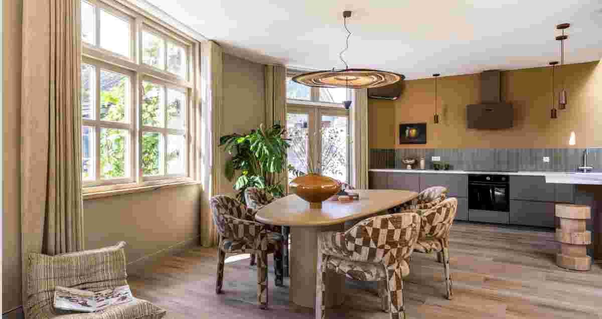 Moderne open woonruimte met eethoek en keuken in warme aardtinten, grote ramen en rieten hanglamp boven tafel.
