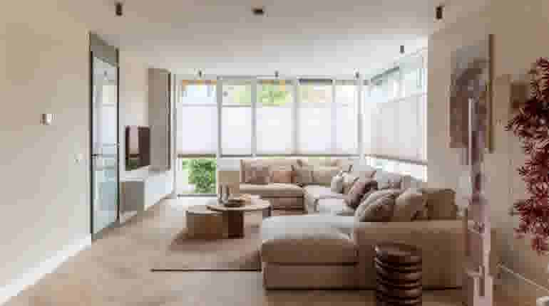 Moderne woonkamer met beige hoekbank, grote ramen met half-transparante gordijnen en decoratieve rode plant.
