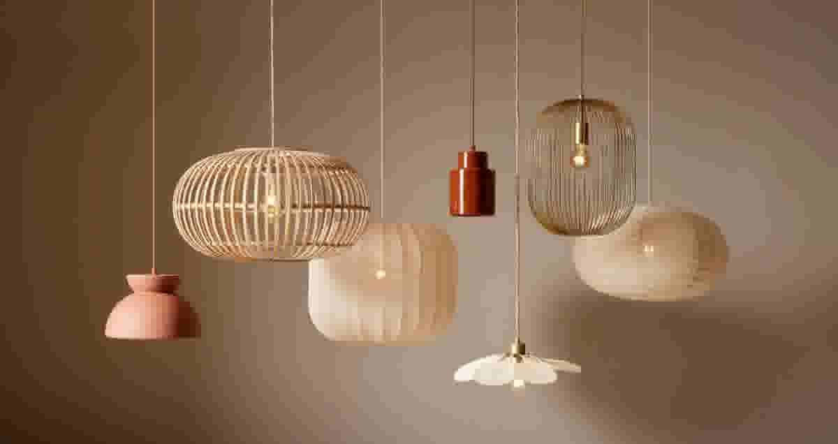 Zes hangende designlampen in warme tinten: roze, bamboe, crème, rood, goud en naturel, tegen een beige achtergrond.