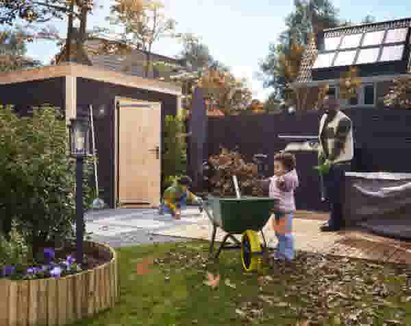 Een man en twee kinderen ruimen herfstbladeren op in de tuin. Een kind duwt een groene kruiwagen, terwijl de vader toekijkt.