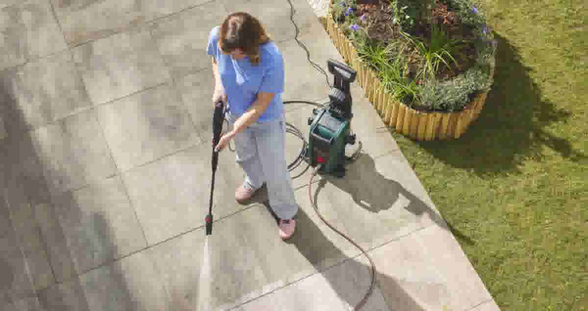 Vrouw in blauw shirt reinigt tegels met een groene hogedrukreiniger in een tuin met bloembak en gazon.
