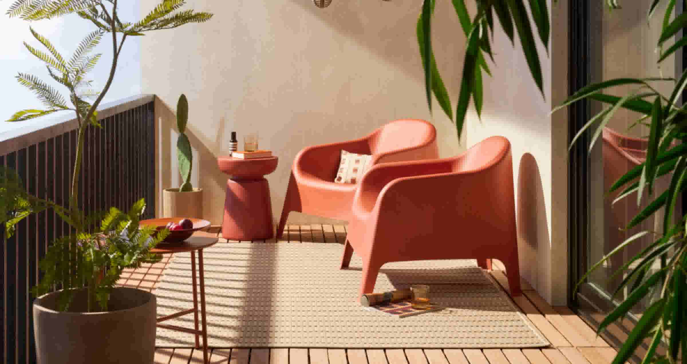 Zonnig balkon met twee koraalrode fauteuils op een crème tapijt, omringd door groene planten en bijzettafels.