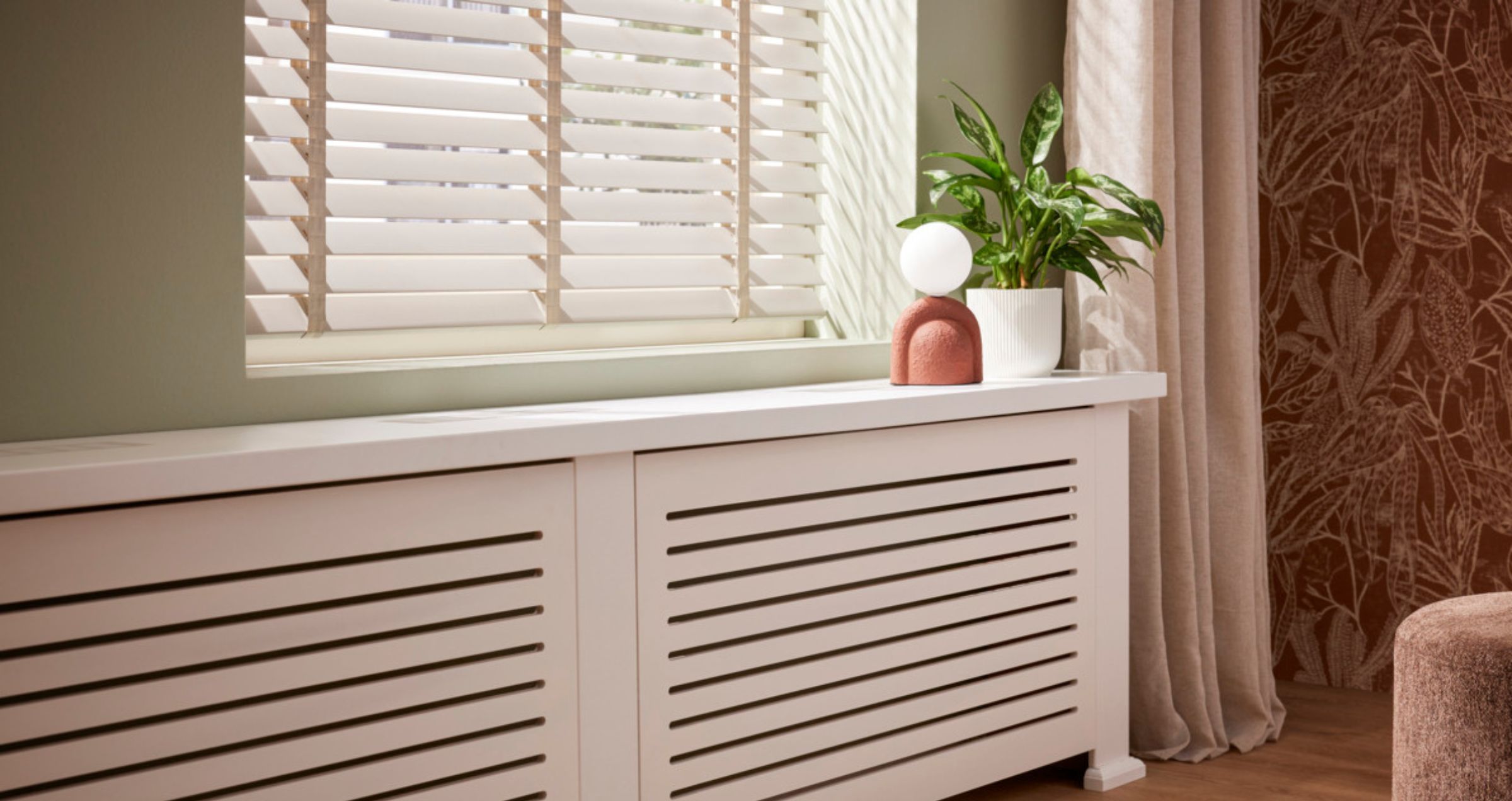 Witte radiatorombouw onder raam met jaloezieën, versierd met een groene plant en terracotta decoratie met witte bol.