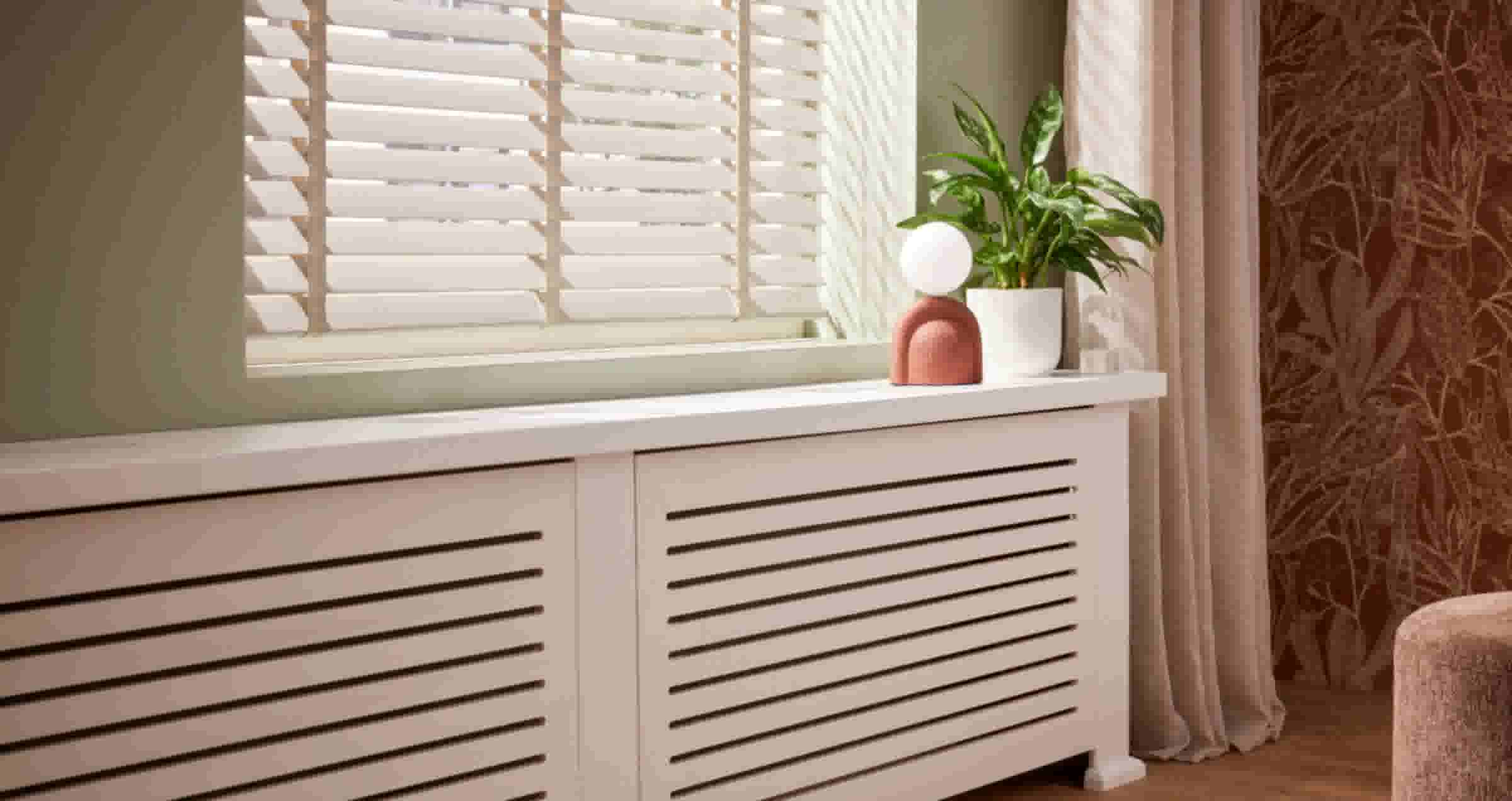 Witte radiatorombouw onder raam met jaloezieën, versierd met een groene plant en terracotta decoratie met witte bol.