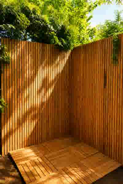 Houten schutting met verticale latten in een hoek, houten tegelvloer, groene bomen en blauwe lucht op de achtergrond.