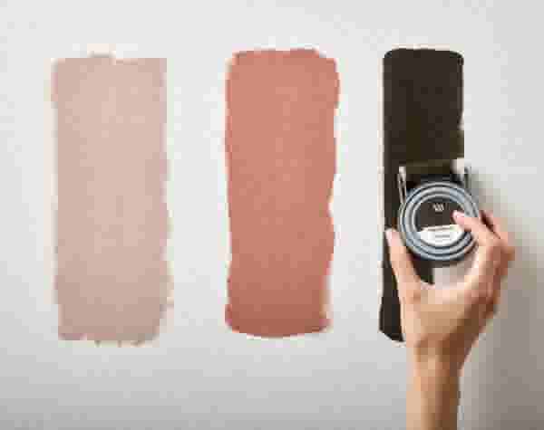 Hand schildert drie verfstrepen op een witte muur: lichtbeige, terracotta en donkerbruin, met een kleurtester in de hand.