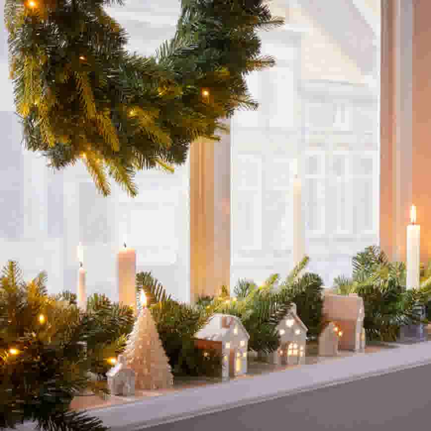 Gezellige kerstdecoratie op vensterbank met verlichte miniatuurhuisjes, kaarsen en dennentakken onder een hangend kersttakje.
