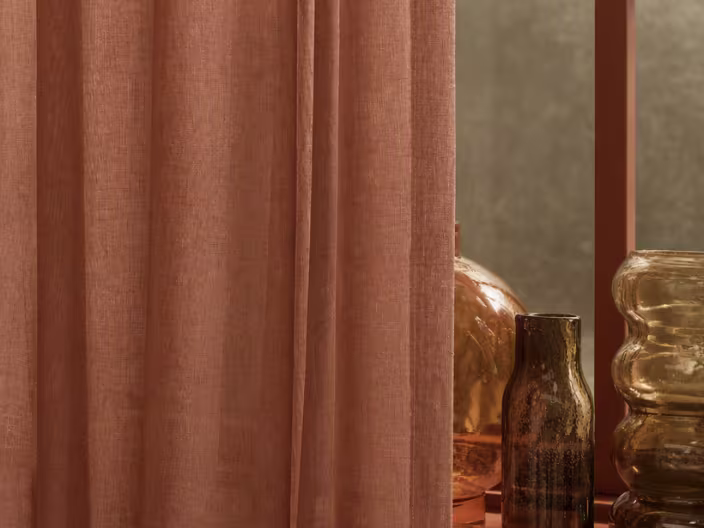 Terracotta gordijn met zachte plooien naast decoratieve bruine glazen vazen en een gouden lamp op een tafel.
