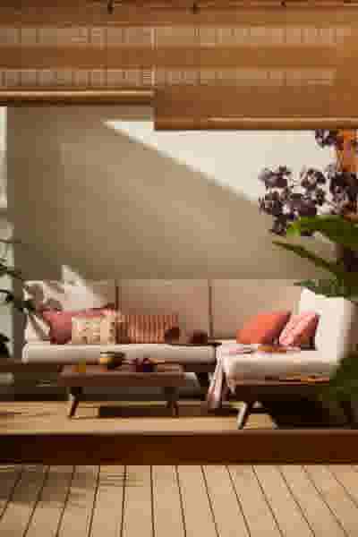 Buitenlounge met cremekleurige hoekbank, roze en oranje kussens, houten salontafel en paarse bloemen. Bamboe rolgordijnen boven.