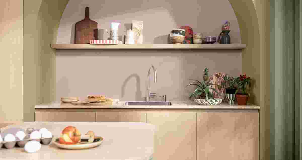 Moderne keuken met roze muur en houten kasten. Plank met keukengerei, gootsteen, planten en fruit op het aanrecht.
