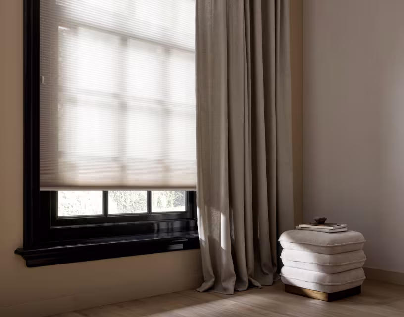 Minimalistisch interieur met zwart raamkozijn, beige plissé gordijn, taupe gordijnen en een witte gestoffeerde poef.