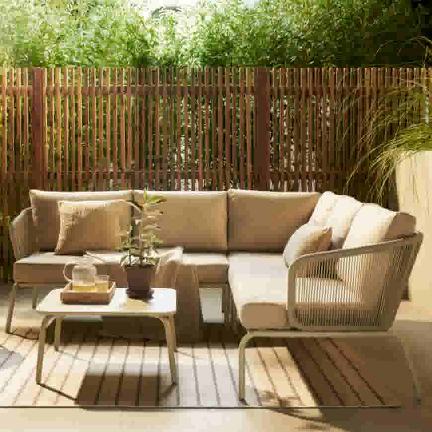 Beige hoekbank op terras met houten schutting, lage tafel met plant en drankje, en siergrassen op de achtergrond.