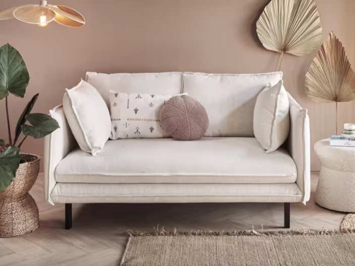 Crèmekleurige tweezitsbank met sierkussens in een warme woonkamer met roze muur, plant en droogbloemen decoratie.