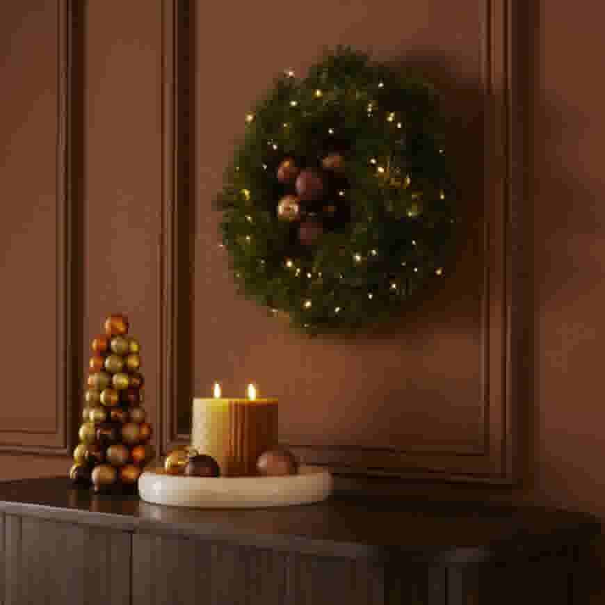 Feestelijke kerstkrans met lichtjes aan bruine muur, eronder een dressoir met kaarsen en een kleine kerstboompyramide.