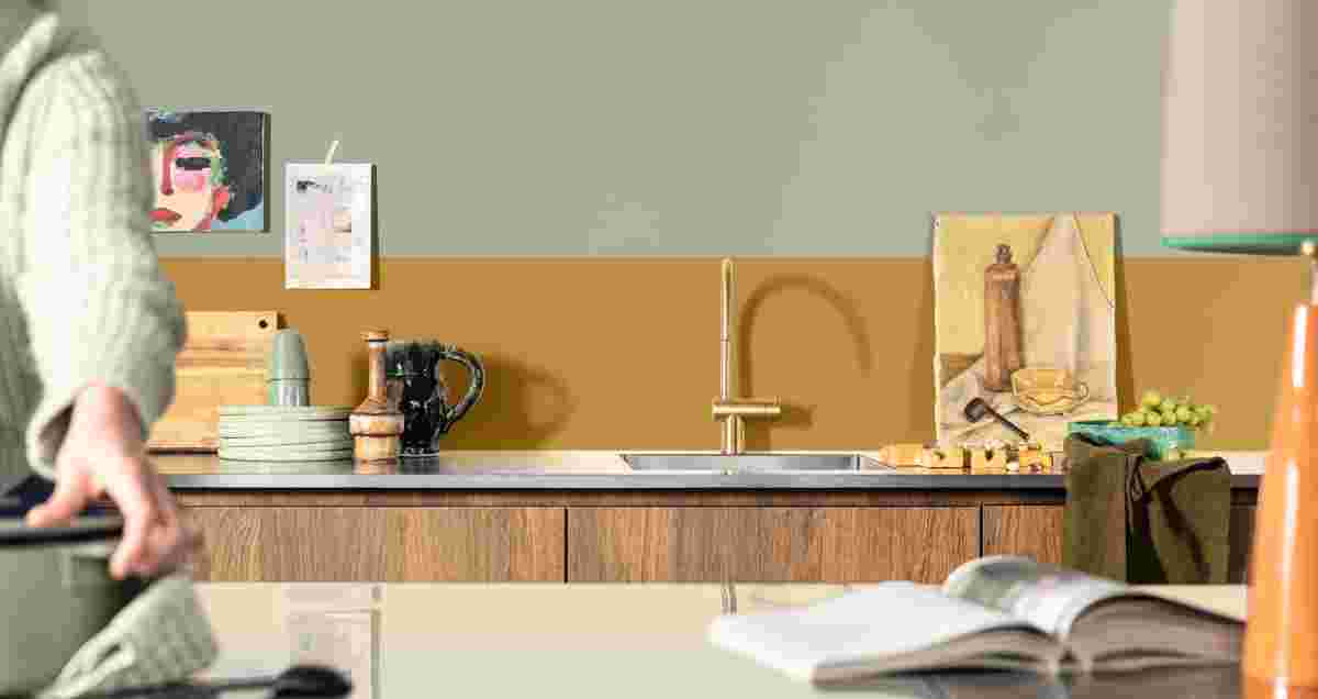 Keuken met oker en salie groene muur, houten kasten, gouden kraan, keramiek, kunstwerken en druiven. Persoon in trui pakt iets vast.